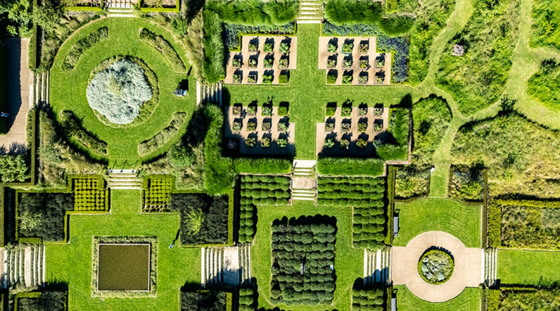 Aerial view of formal hotel gardens with geometric hedges, circular lawns, raised planting beds and winding pathways.
