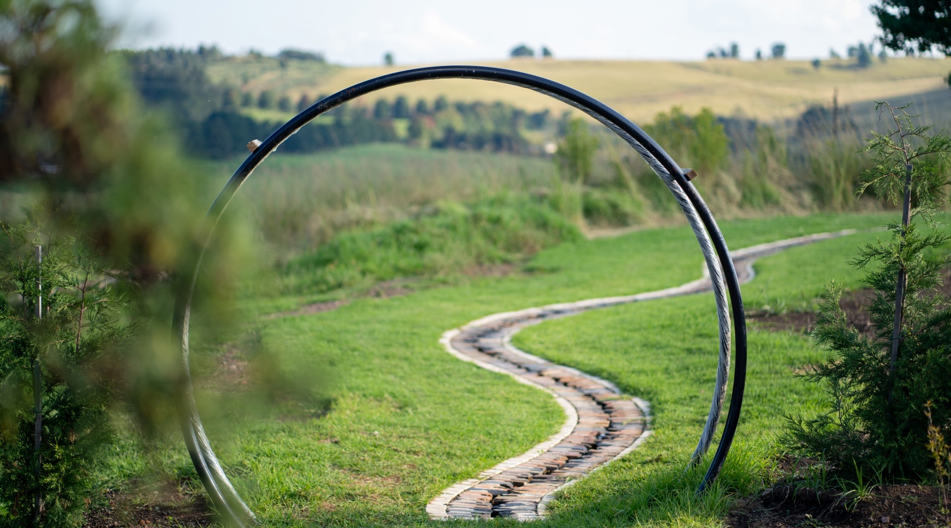 Winding stone garden path framed by a circular metal arch, manicured lawn and young trees with rolling countryside views.