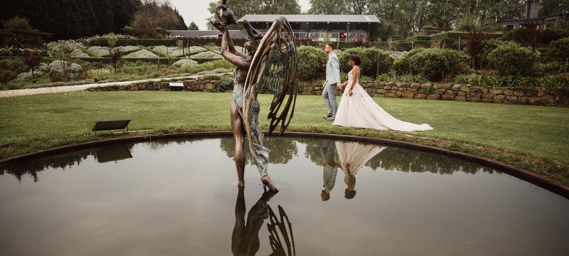Bronze winged statue reflected in a pond, bride in flowing gown and groom walking hand-in-hand across manicured garden wedding venue.
