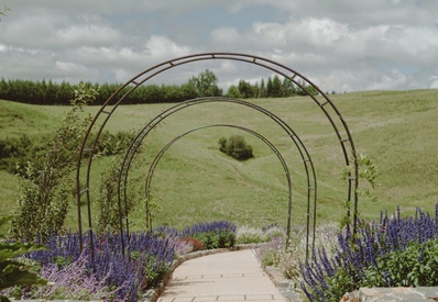 Garden walkway framed by metal arched trellises, stone walls and beds of purple flowers with rolling green hills.