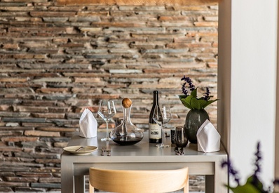 Intimate table for two with wine decanter, bottle and glasses, folded napkins and floral vase against textured stone wall.