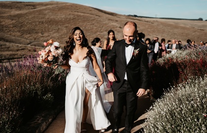 Newlyweds walk a wildflower-lined path after an outdoor ceremony on a sunlit hillside; bride in white gown with bouquet, groom in black tux