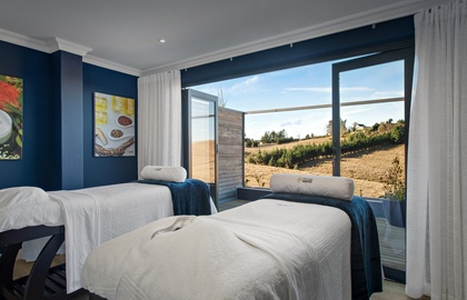 Dual spa massage beds with white linens and bolster pillows in a navy treatment room opening to a sunlit hillside deck view.