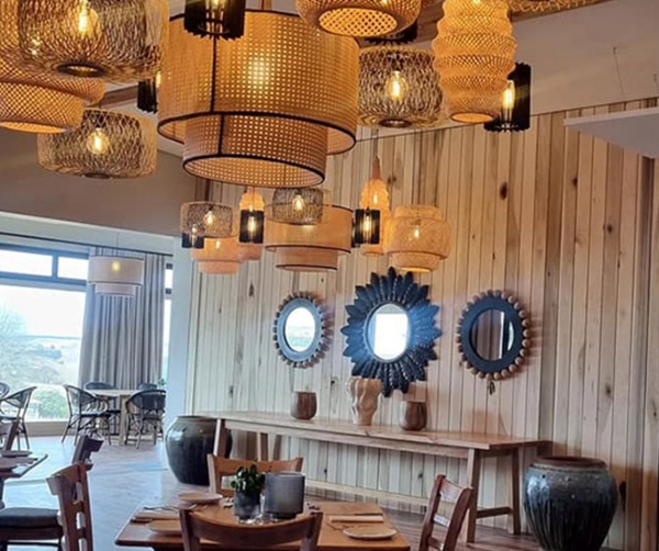 Warm dining room with wood-paneled wall, woven pendant lights, decorative mirrors, set tables and large windows.