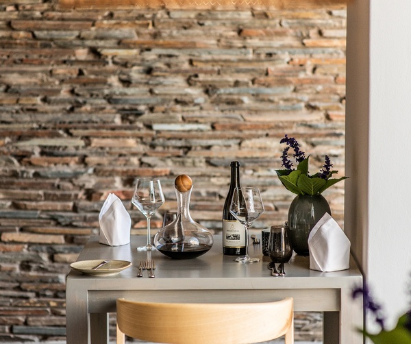 Intimate hotel dining table with wine bottle, decanter, wine glasses, napkins and purple flowers by a stacked stone wall