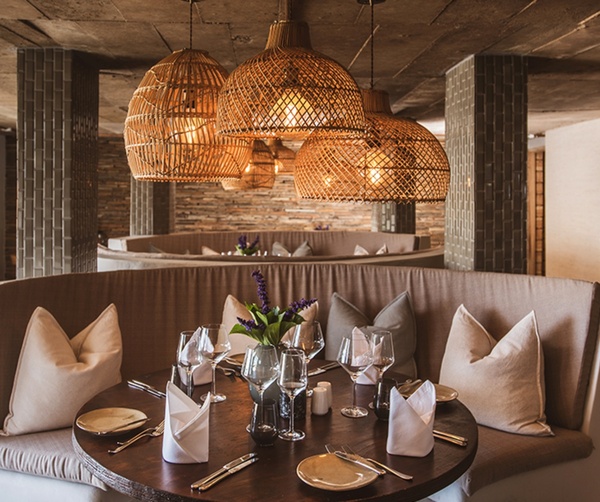 Cozy hotel restaurant with curved banquette seating, plush cushions, round wood table set for dinner and warm woven pendant lights.