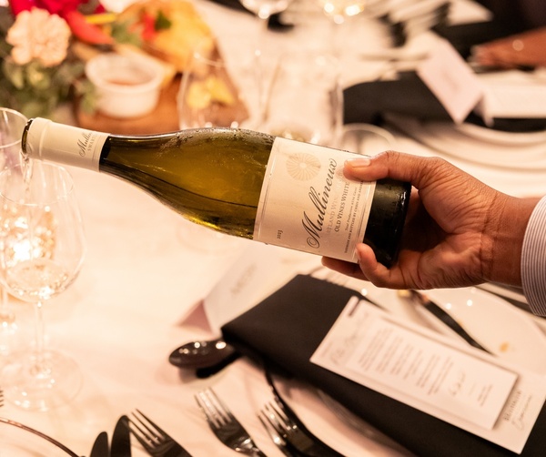 Server pouring chilled white wine into glass at a candlelit, floral-decorated dinner table with menus and polished cutlery.