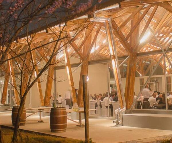 Warmly lit timber pavilion with vaulted wooden beams and glass walls, guests dining at banquet tables inside; outdoor barrel tables.