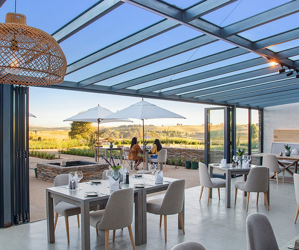 Open-air restaurant with glass roof, wicker pendant lights and patio umbrellas, dining tables overlooking rolling countryside