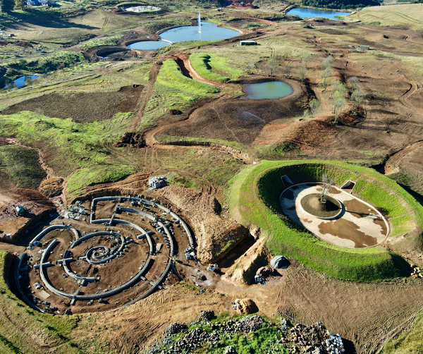 Aerial view of landscaped grounds: circular earth-sheltered courtyard with tree, spiral foundation, ponds and rolling grassy hills.