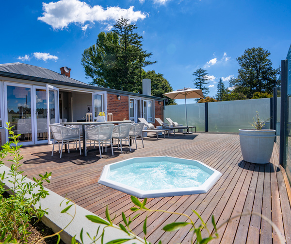 Sunlit private deck with bubbling hot tub, dining table with wicker chairs, sun loungers and umbrella, frosted glass panels.