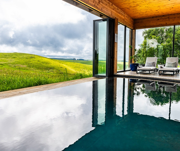 Infinity pool with reflective water beside a wooden spa pavilion; reclining lounge chairs and glass walls open to green hills.