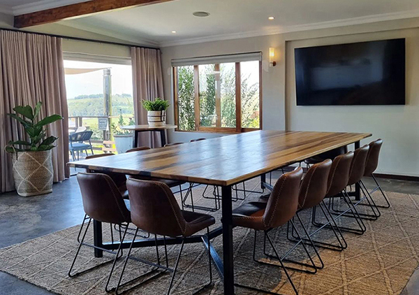 Boardroom-style meeting room with long wooden table, leather chairs, wall TV, exposed beam, woven rug and terrace views