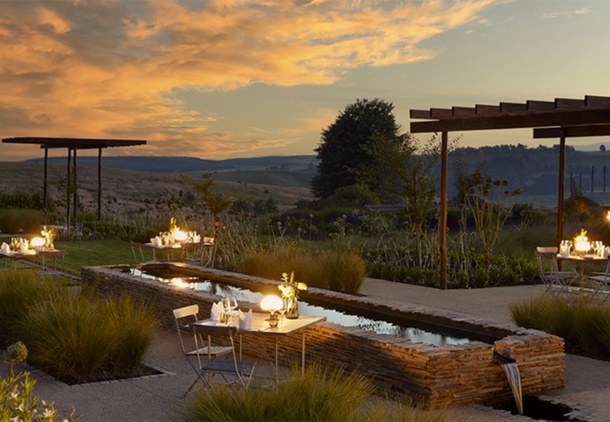 Outdoor candlelit dining by a stone water channel, pergolas and landscaped terraces with rolling hills at sunset.