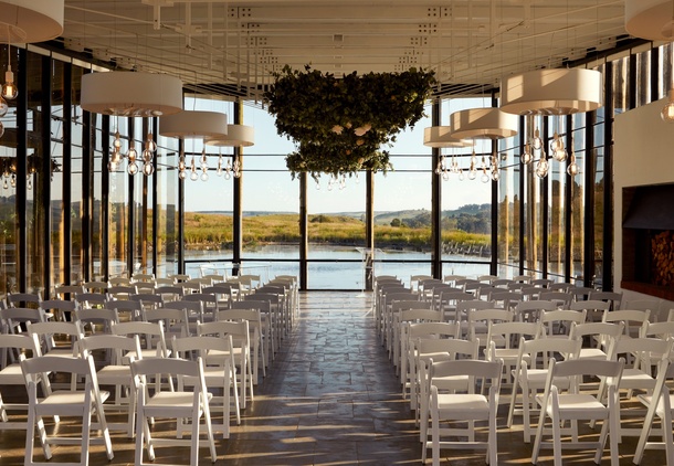 Glass-walled waterfront ceremony room with rows of white folding chairs, hanging drum lamps and bulb pendants, greenery chandelier.