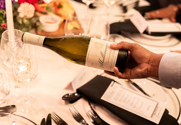 Server pouring chilled white wine into glass at a candlelit, floral-decorated dinner table with menus and polished cutlery.