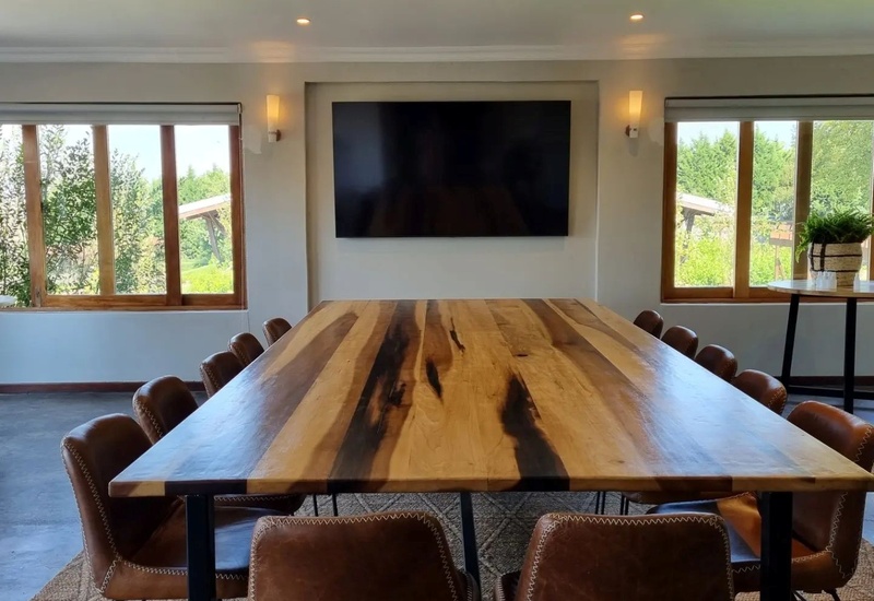 Large rustic conference room with long wooden table, leather chairs, wall-mounted TV, windows overlooking lush garden.