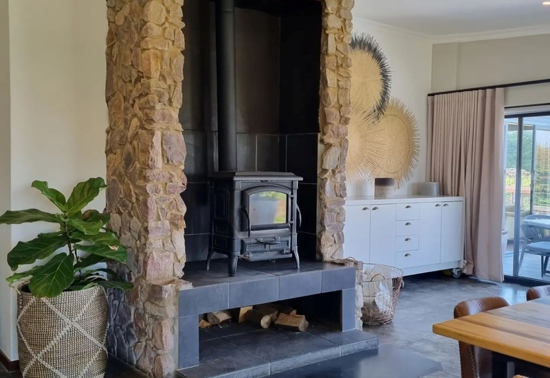 Cozy lounge with stone-framed wood-burning stove, stacked firewood, woven plant basket, white sideboard and sliding doors to sunlit terrace.