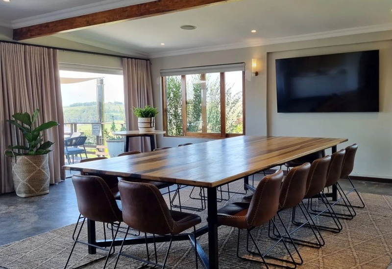 Long wooden conference table with leather chairs, wall TV, potted plants and sliding doors to a terrace with rolling countryside views