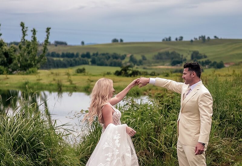 Bride twirling in lace gown with groom in cream suit on lakeside lawn, rolling green hills and cloudy sky backdrop
