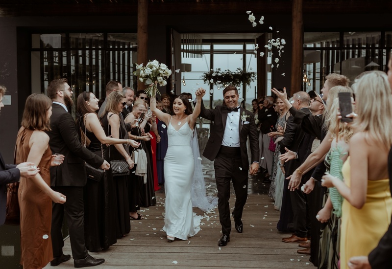 Newlyweds exit a hotel ceremony on a wooden deck beneath a covered terrace; bride holds bouquet, guests in formalwear toss white petals.