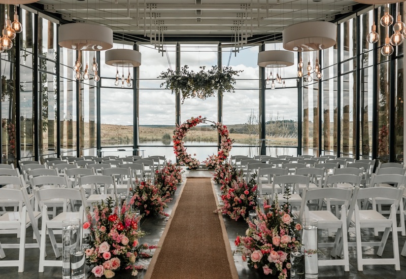 Glass-walled lakeside ceremony space with white chairs, floral-lined aisle and circular flower arch under pendant lights