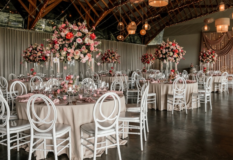 Banquet hall set for wedding with round tables, blush linens, tall pink floral centerpieces, white chairs and warm hanging lights.