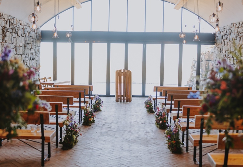 Stone-walled chapel with arched glass wall, wooden benches, floral-lined aisle, central wooden lectern and warm hanging pendant lights