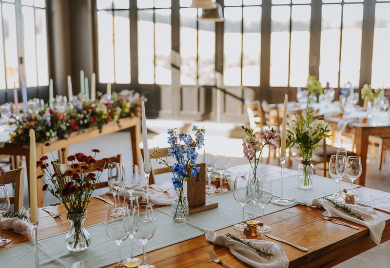Sunlit dining room with wooden banquet tables set for celebration, glassware, taper candles and fresh floral centerpieces.