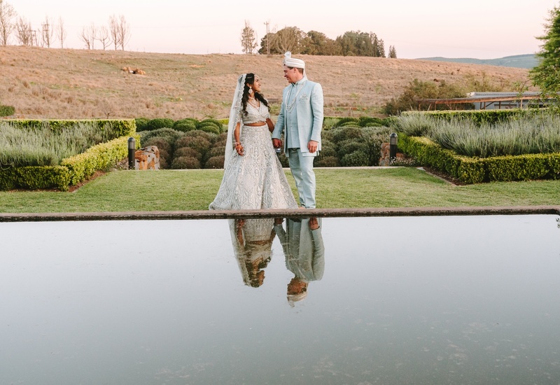 Bride and groom in ornate traditional attire holding hands on manicured lawn by a reflecting pool at a countryside estate.