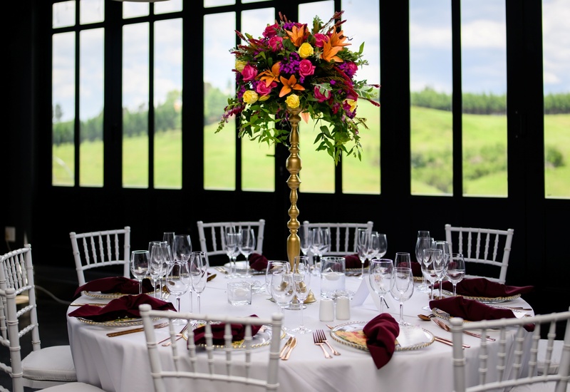 Elegant banquet table with gold floral centerpiece, burgundy napkins, glassware and white chiavari chairs by green fields