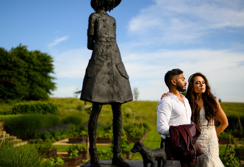 Bride in lace gown and groom holding a jacket pose beside bronze girl-and-dog sculpture in sunlit hotel garden terrace