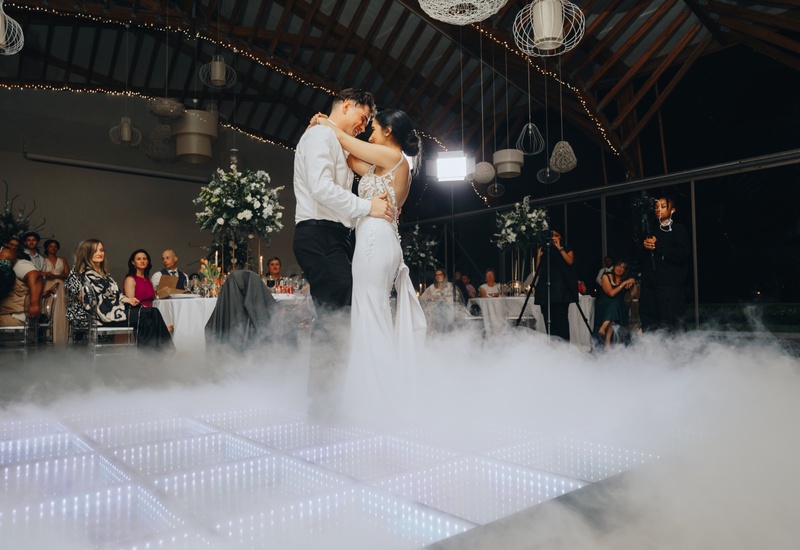 Bride and groom share first dance on illuminated LED floor amid low fog, string lights and floral-tabled wedding reception.