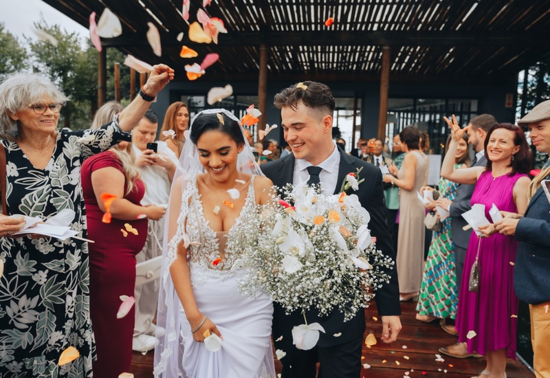 Bride in lace gown and veil holding large white-orchid bouquet, groom in suit, guests tossing rose petals on a hotel terrace under a wooden pergola.