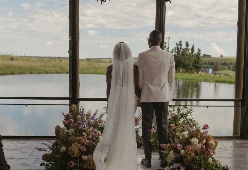 Bride and groom stand hand-in-hand before floor-to-ceiling windows overlooking a lake, surrounded by flowers, candles and hanging greenery.