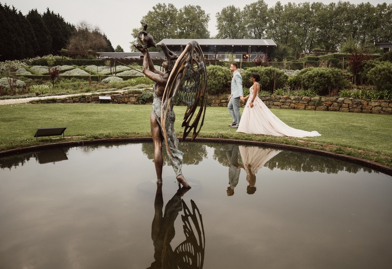 Bronze winged statue reflected in a pond, bride in flowing gown and groom walking hand-in-hand across manicured garden wedding venue.