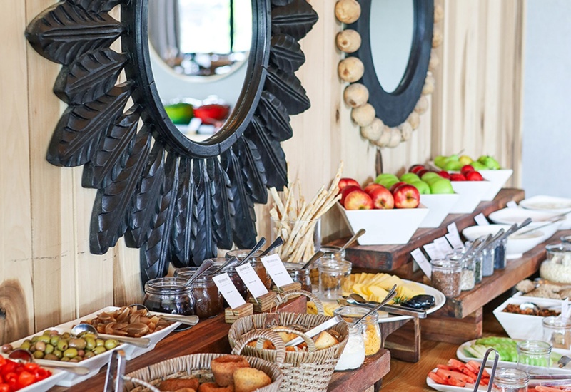 Rustic breakfast buffet with sliced breads, wicker baskets, jams, bowls of fresh fruit and decorative round wooden mirrors.