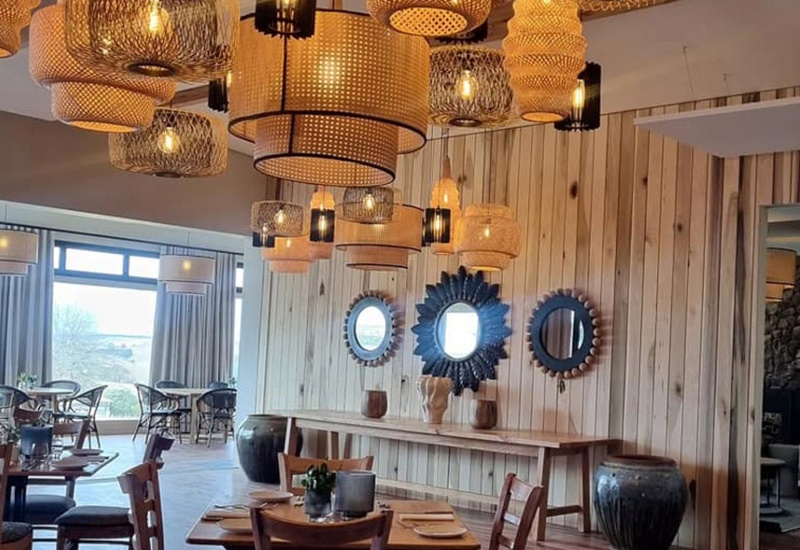 Cozy hotel dining room with clustered woven pendant lights, wood-paneled wall with round mirrors, set tables and large windows.