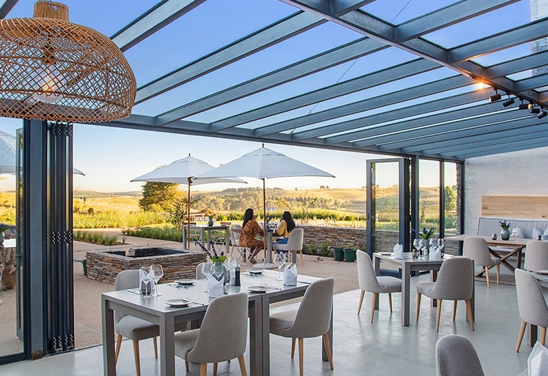 Covered glass-roof dining terrace with wicker pendant lamps, set tables, two guests under umbrellas and rolling countryside views.
