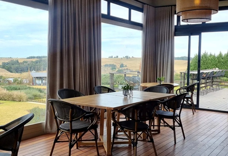 Wood-furnished dining area with woven chairs and glass walls opening to a deck with bar seating and rolling hill views