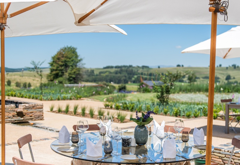 Alfresco dining table set with glassware and napkins under white umbrellas, overlooking landscaped garden and rolling countryside.