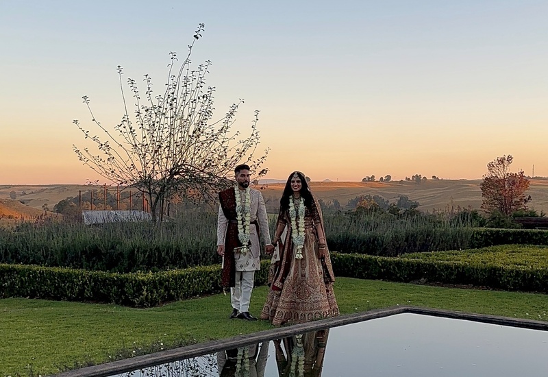 Newlyweds in traditional South Asian wedding attire strolling beside a reflective pool in manicured gardens at sunset