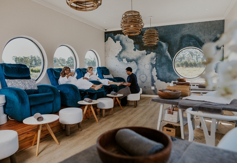 Plush blue pedicure chairs by round windows, rattan pendant lights, guests in robes sip drinks while receiving pedicures.