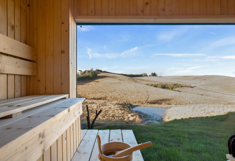 Wood-lined sauna with bench, bucket and ladle, floor-to-ceiling window framing rolling golden hills, pond and grassy countryside view.