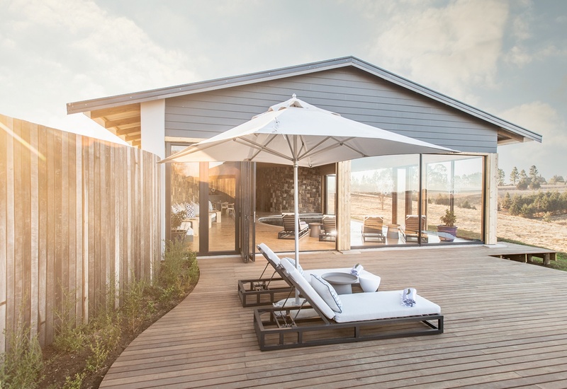 Private wooden deck with cushioned sun loungers and large parasol, sliding glass doors to a modern villa and countryside views.