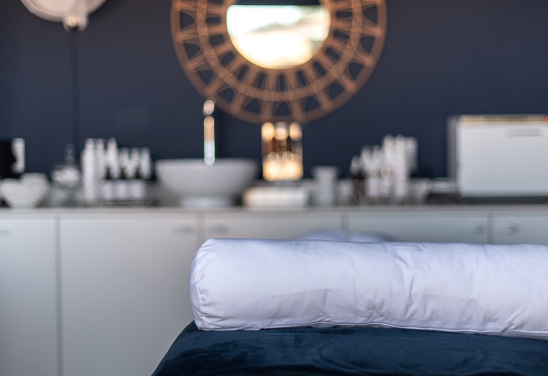 Spa treatment table with white bolster pillow on navy cover; blurred sink, skincare bottles and decorative round mirror.