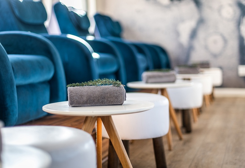 Plush teal pedicure chairs in a spa with white footstools and round tables holding rolled gray towels and greenery.