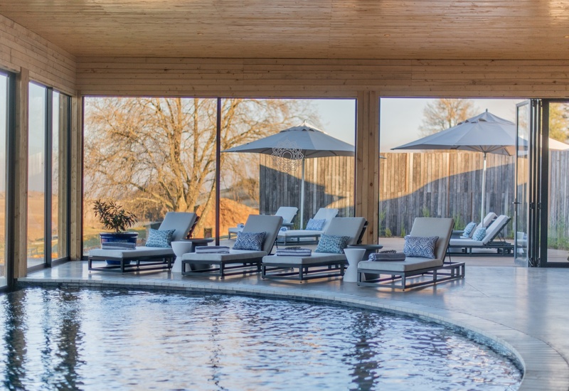 Indoor heated pool with curved edge, cushioned lounge chairs and umbrellas by floor-to-ceiling glass overlooking countryside views.