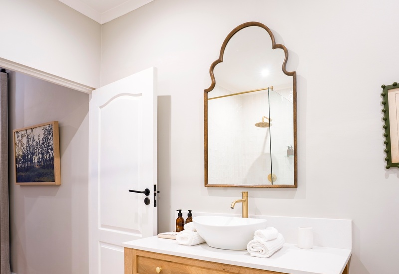Bright bathroom with wooden vanity and vessel sink, brass faucet, arched decorative mirror, glass rainfall shower, rolled towels.