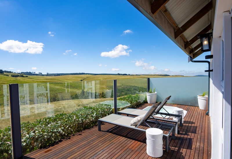 Private wooden terrace with two sun loungers, glass railing and potted plants overlooking rolling green hills under blue sky.